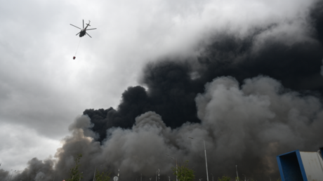 A firefighting helicopter is carrying water as black smoke billows from the area of Gazprom Neftas Moscow oil refinery, Moscow, June 18, 2026.